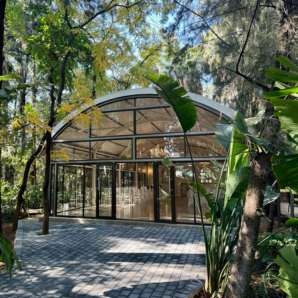 Curved glass entrance of the Glass Dome Chapel surrounded by mature trees and tropical foliage